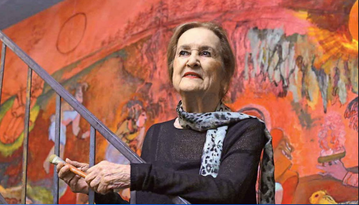 Rina Lazo, de unos 80 años, de pie en una escalera metálica delante de un mural. Sostiene un pincel en la mano.