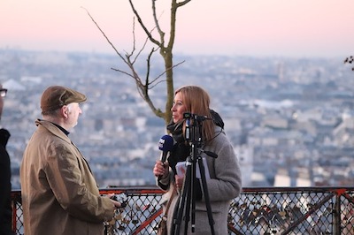 Imagen en exteriores. Una mujer de abrigo con un micrófono en la mano entrevista a un hombre. Panorámica de una ciudad al fondo.
