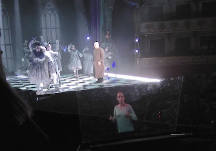 Scene of The Addams Family in the theater. Like floating in the air, on the transparent lectern the sign language interpreter is seen. The interpreter raises her left arm and has her right arm folded against her body. 