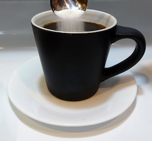A cup of black coffee. White sugar falls in the coffee from a spoon.