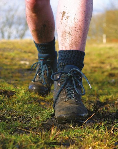 Legs of a man in hiking boots walking on a field of grass. The legs are spattered with mud.