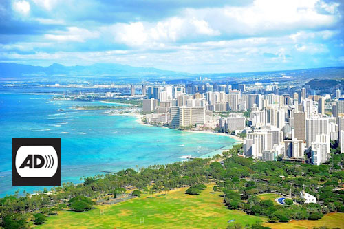 Vista panorámica de Honolulu. El mar turquesa baña las playas de la ciudad, junto a cuya costa se levantan un gran número de edificios altos. Sobre la imagen, en la esquina inferior izquierda, aparecen las letras AD en negro en un fondo blanco. 