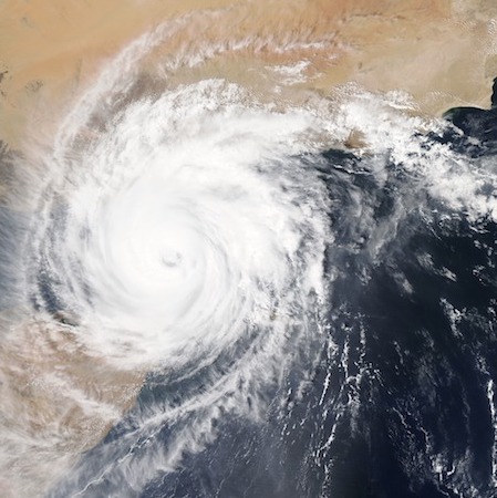 Imagen satelital. Un huracán gira sobre una costa. El huracán es una gran nube blanca en forma de espiral que tiene largo brazos. El centro de la espiral es denso y a medida que se alejan del centro, los brazos son menos densos.