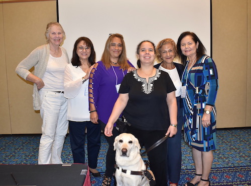 Indoors. Six women smiling stand and pose standing next to each other. Judy Matthews, with Keats, her service dog, stands toward the center, in front of the other women.