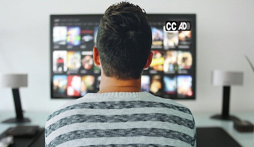 Un hombre sentado frente a una pantalla de televisión. En la esquina superior derecha de la pantalla de televisión están los íconos de closed caption y audio descripción.