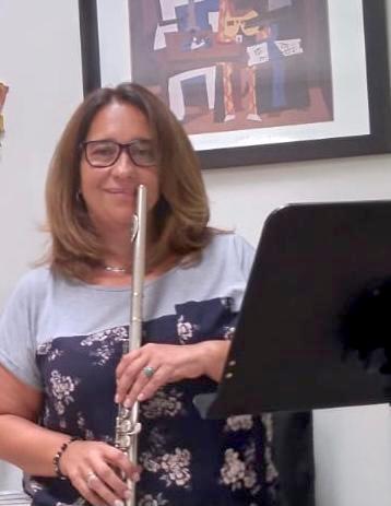 En un salón, Maria Victoria Diaz, sostiene una flauta parada junto a un atril.