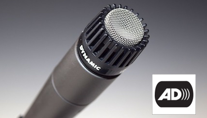 Un micrófono apuntando hacia arriba y hacia la derecha. Abajo a la derecha, las letras AD que simbolizan audio descripción.