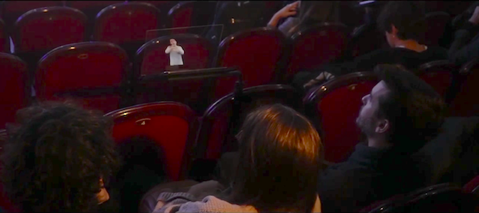 Varias personas sentadas en un teatro observan un atril transparente delante de ellas. El atril muestra una intérprete de señas que viste una blusa azul clara.