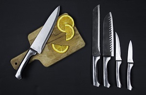Set of 5 stainless steel knives over a black background. One of the knives rest on a wood cutting board, next to 2 orange slices.