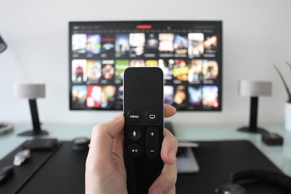 A hand holds a remote control in front of a TV screen that shows a grid of movie pictures.