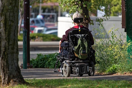 Una persona en una silla de ruedas está en un sendero de cemento frente a una calle.