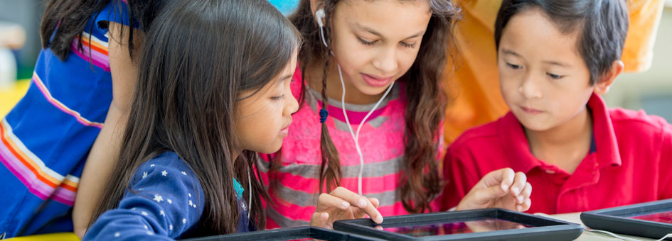 A girl wearing headphones touches the screen of a tablet. 4 children around her look at the screen.
