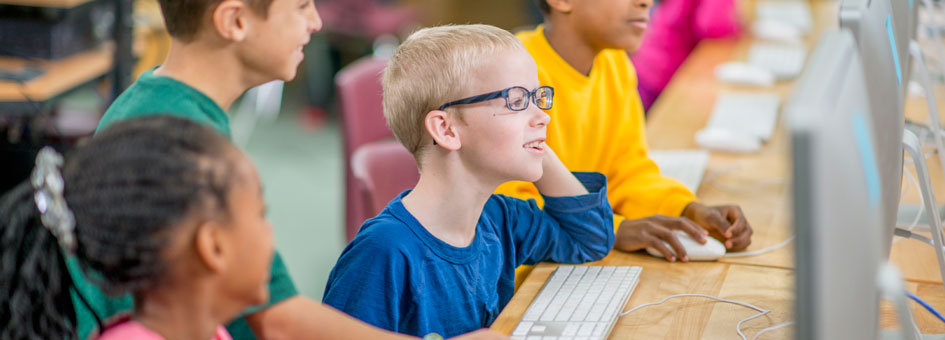 Computer room. A smiling boy with glasses  looks at his screen, another boy standing next to him smiles