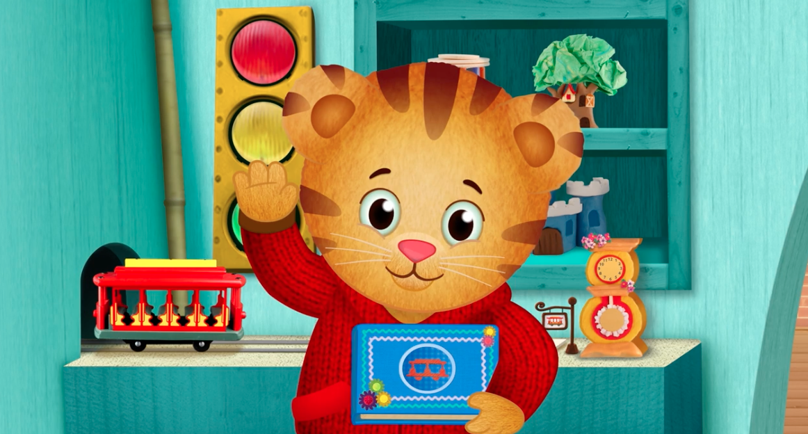 Animated character Daniel Tiger holding a blue book. Toys and a traffic light are visible in the background