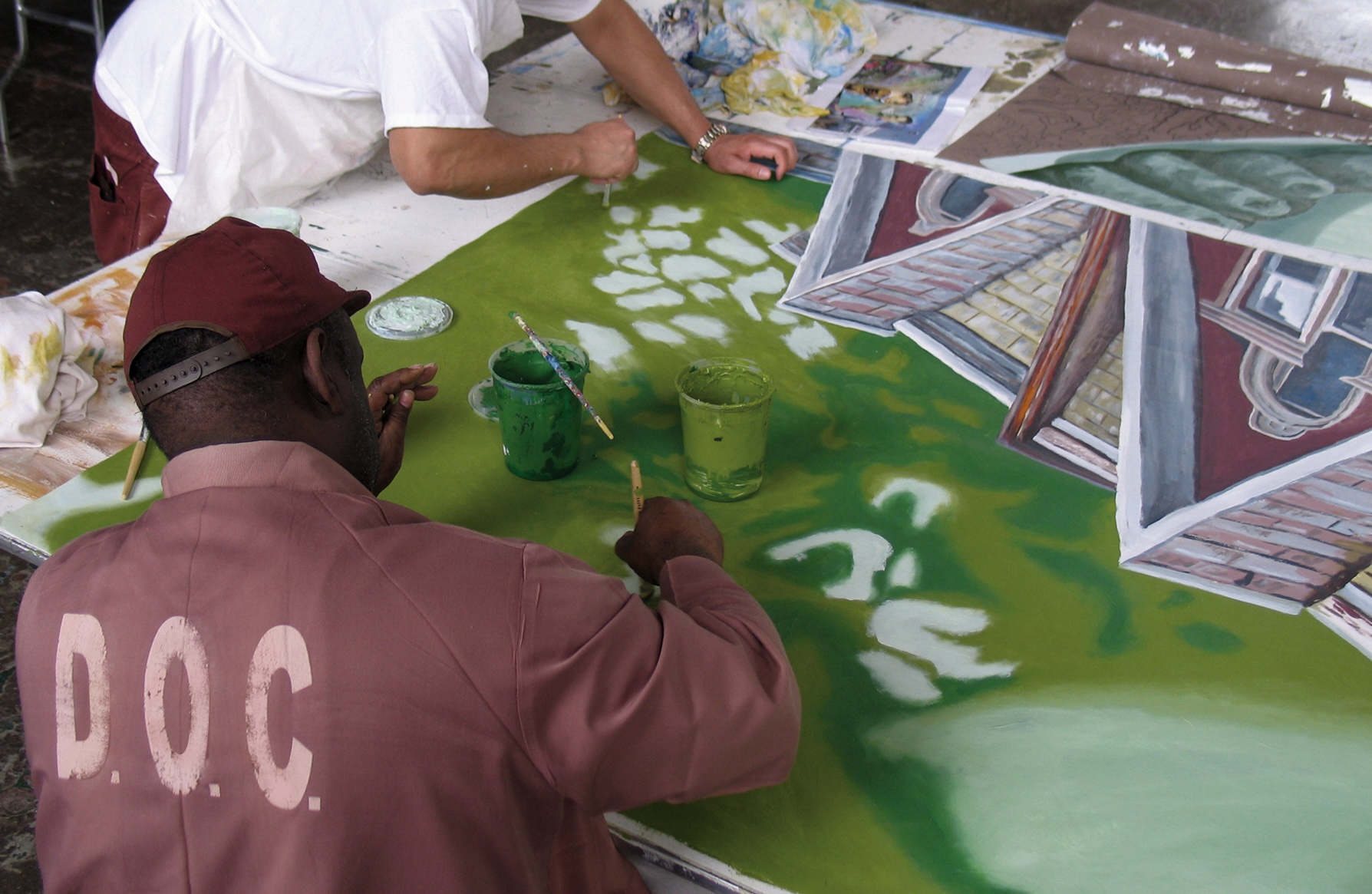 Two men in prison uniforms painting a canvas together on a table