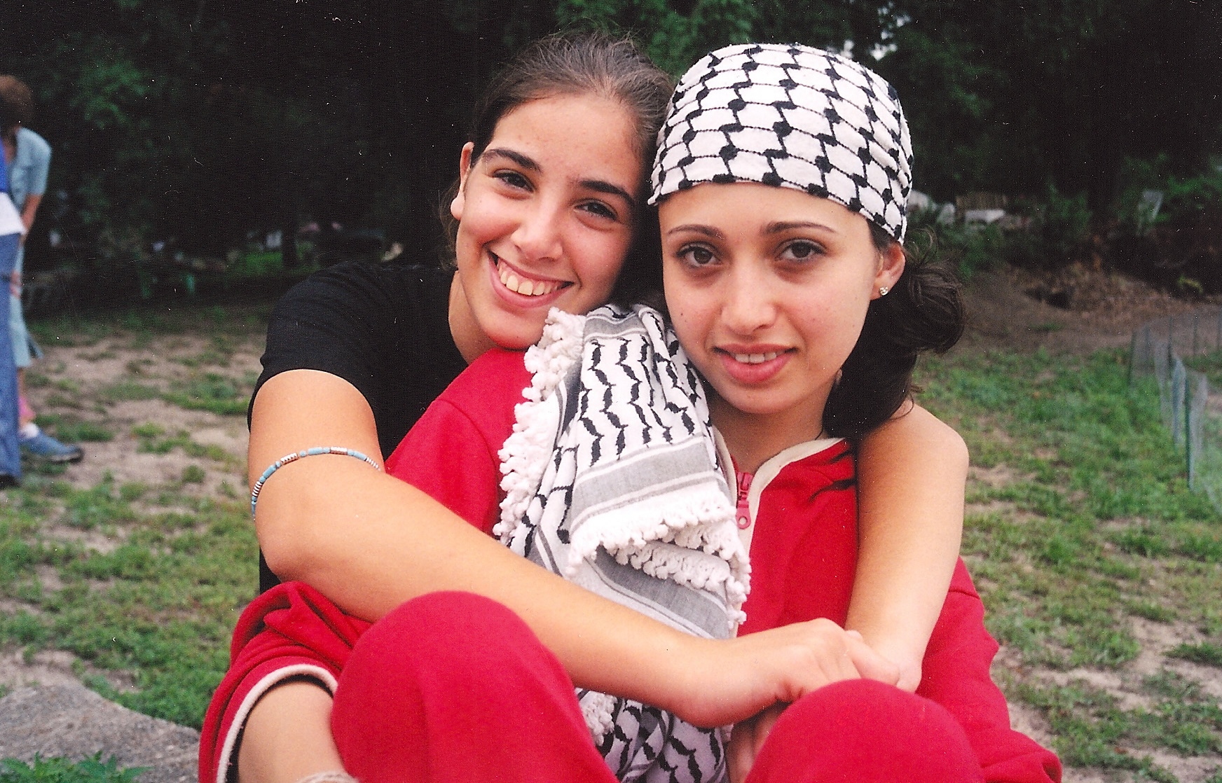Photograph of two young women in a park. One woman smiles and hugs the other, who wears a Palestinian scarf on her head.