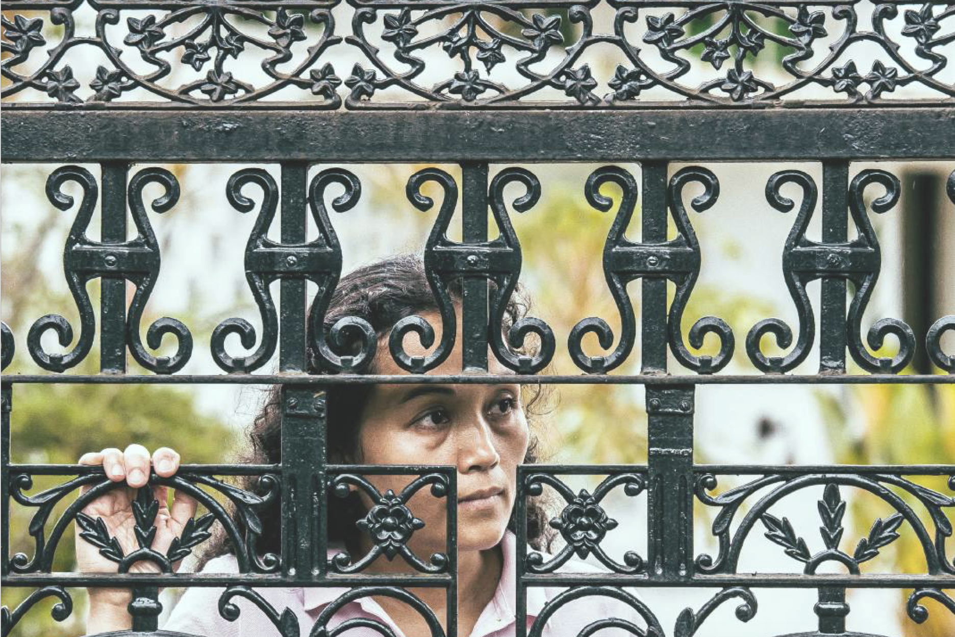 A woman behind a tall, ornate iron fence looks through the bars.