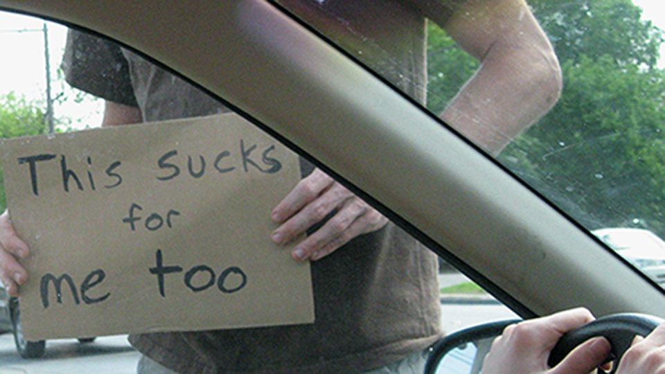 Person holding cardboard sign with text 'This sucks for me too' seen through car window