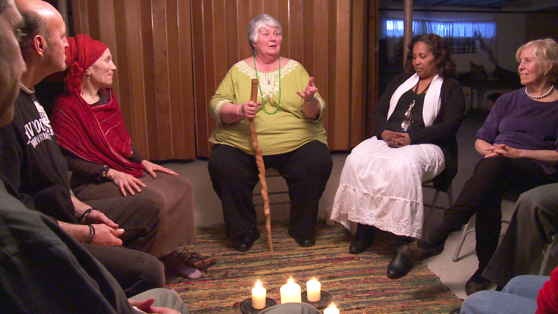 Group of seated adults, mostly women, in a circle. One woman speaks while holding a wooden cane. Lit candles adorn the center