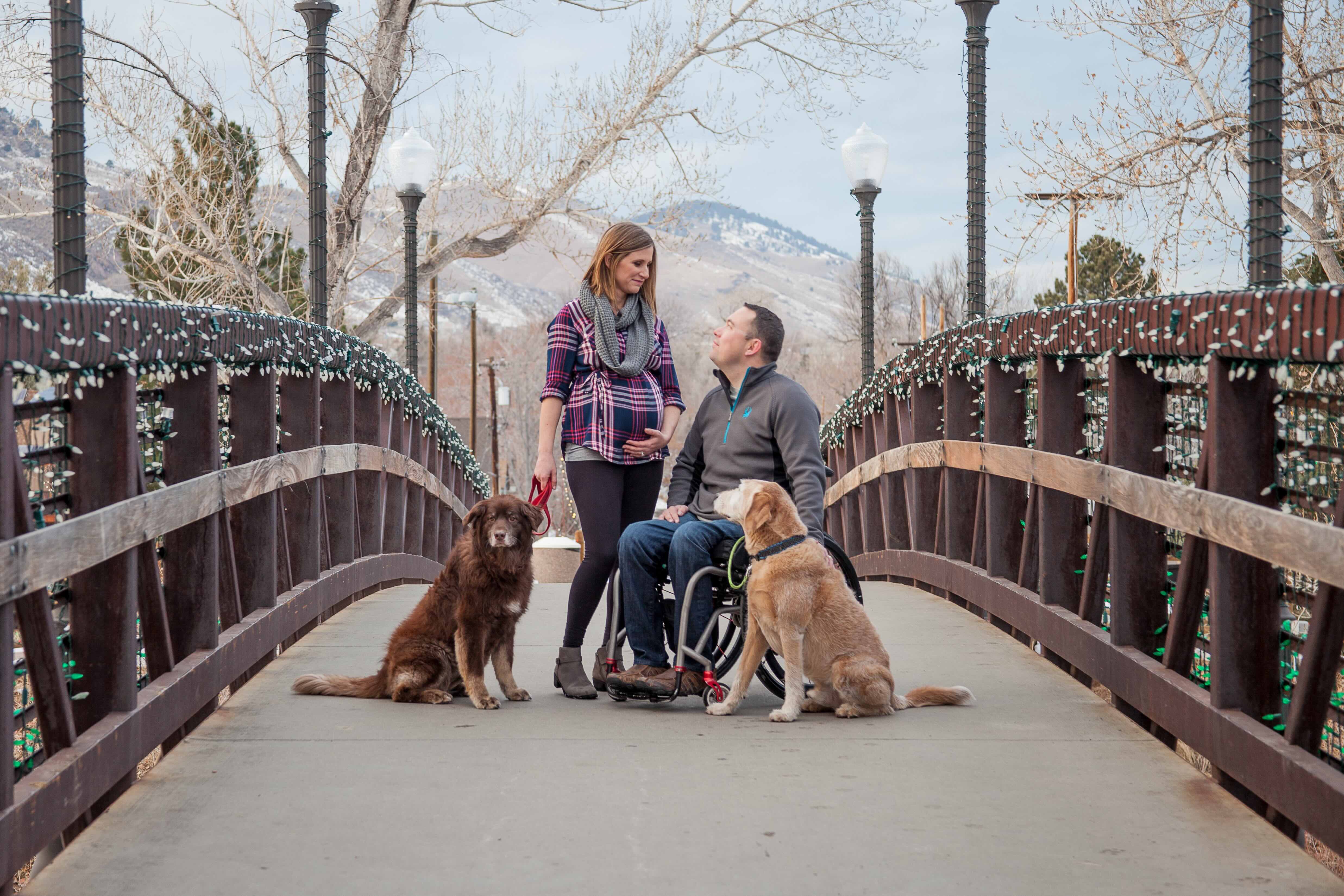 On a wooden bridge, a man in a wheelchair looks at a smiling pregnant woman standing next to him. Next to each one, there is a dog.