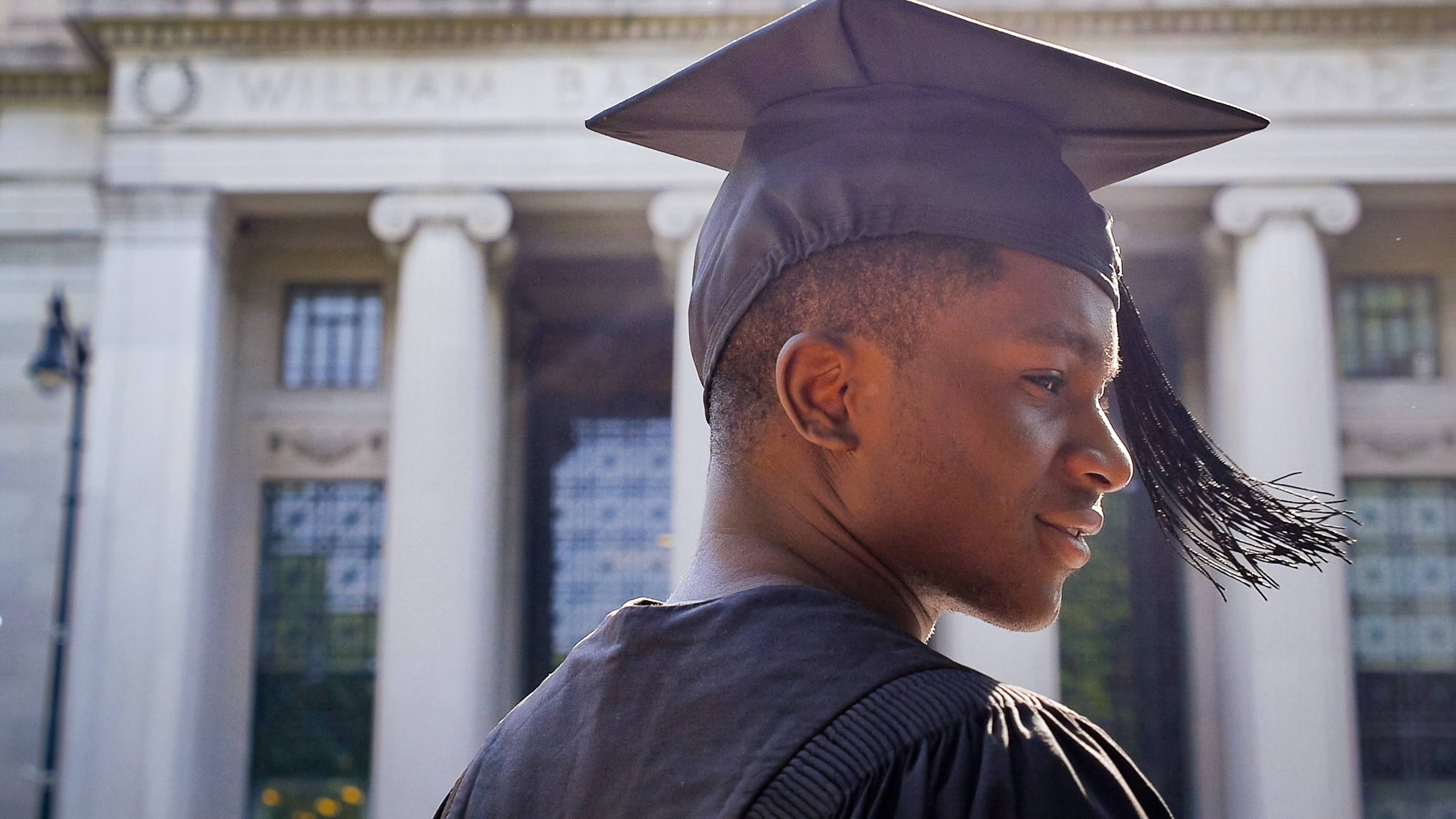 A smiling young black man in graduation cap and gown stands before a tall white stone building with columns.