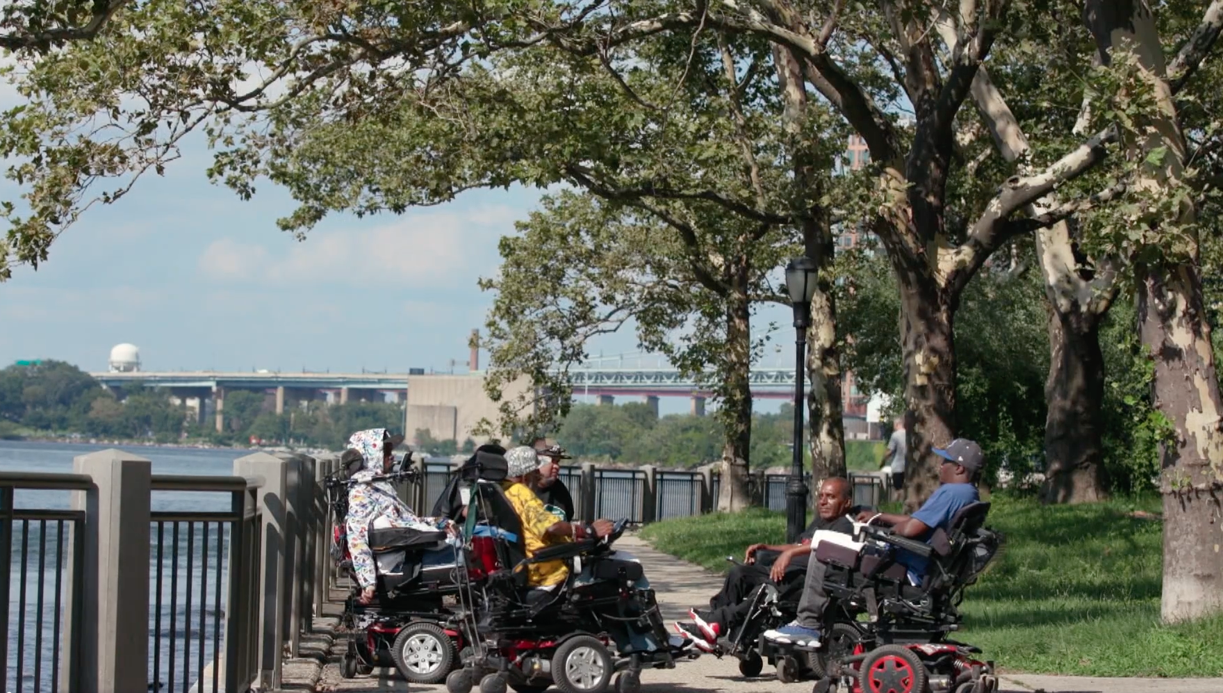 In a park next to a river, six men in powered wheelchairs form a circle.