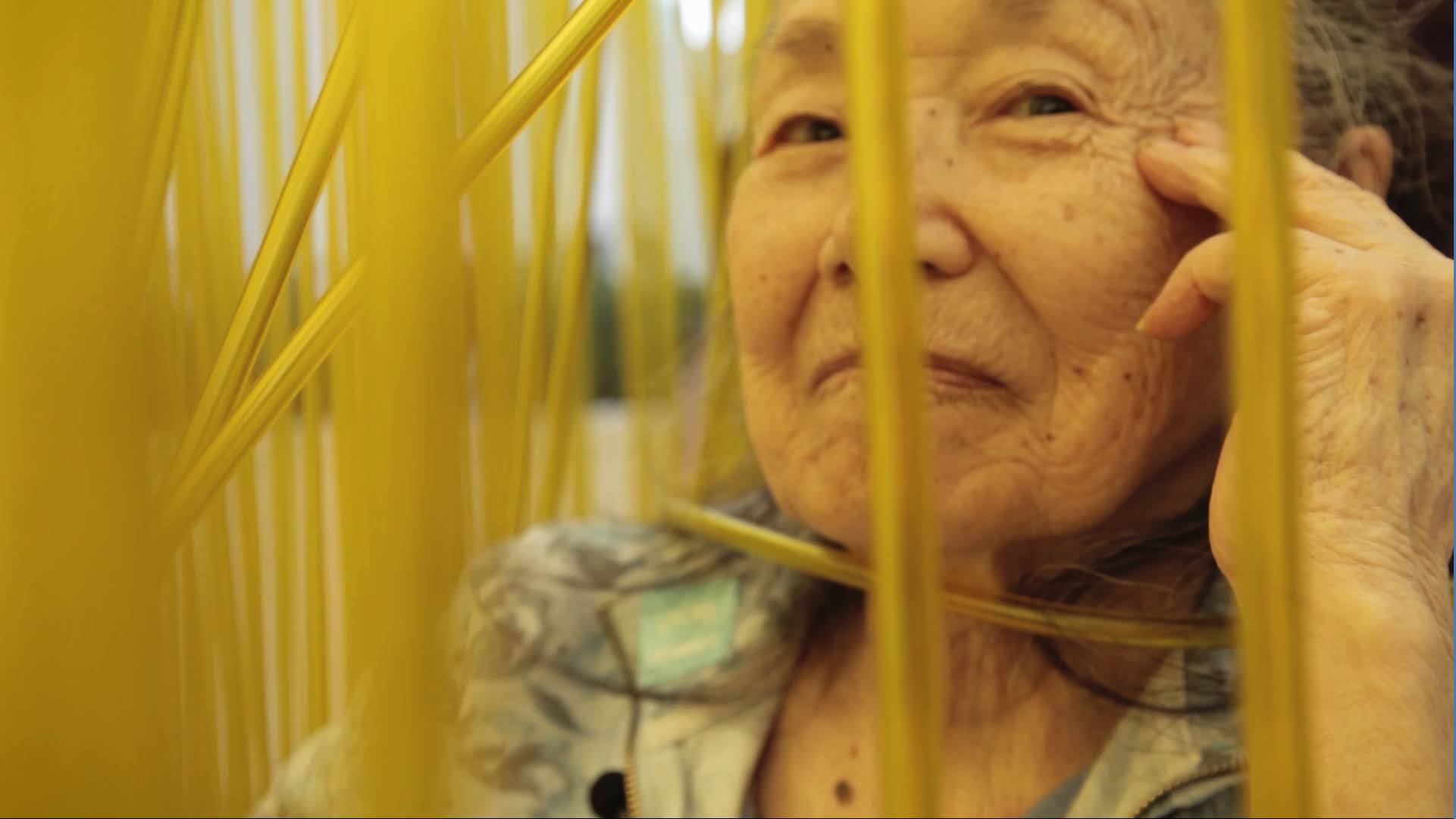 An old woman smiles behind a curtain of yellow plastic strings.