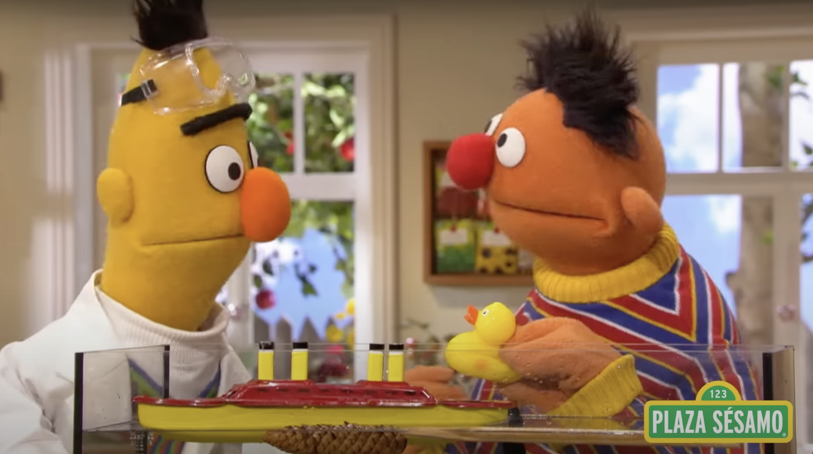 Beto and Enrique from Sesame Street, with Beto wearing a white coat and laboratory glasses. In front of them are a toy boat and a rubber duck