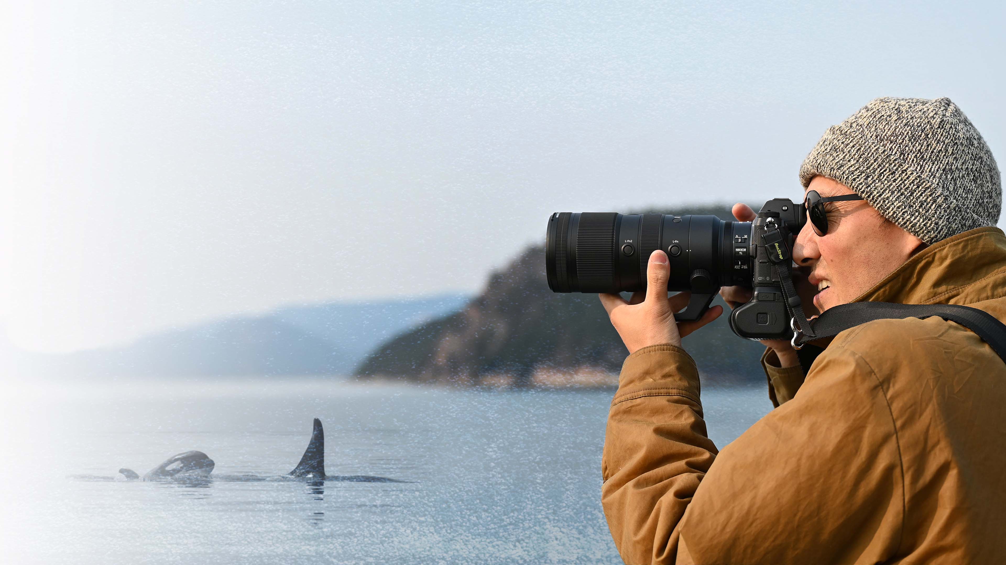 A man in a woolen hat and jacket holds a camera with a long lens and looks through it. A pair of killer whales emerge from the ocean in the background.