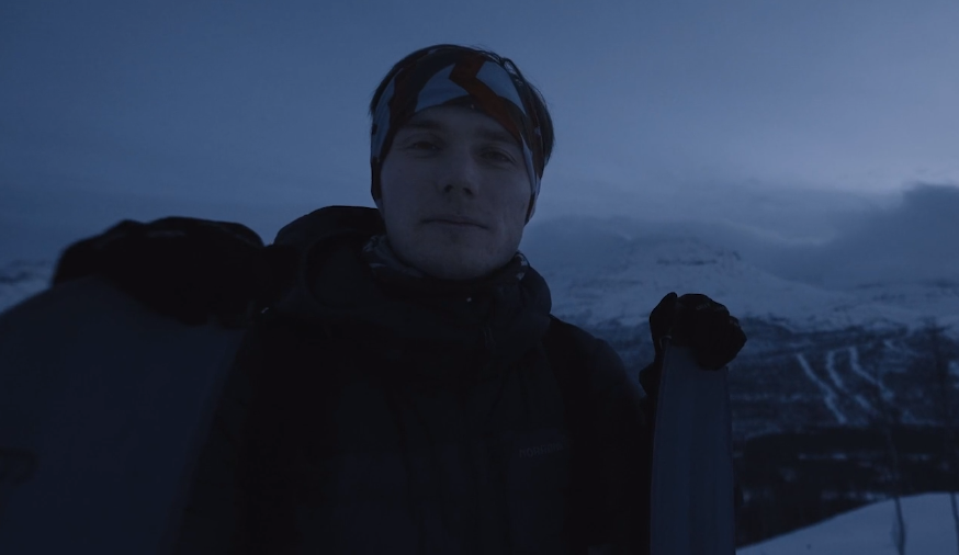 Dimly lit image. A man stands in front of a snow-covered mountain range in the background. He wears a thick winter jacket and has a bandana around his head.