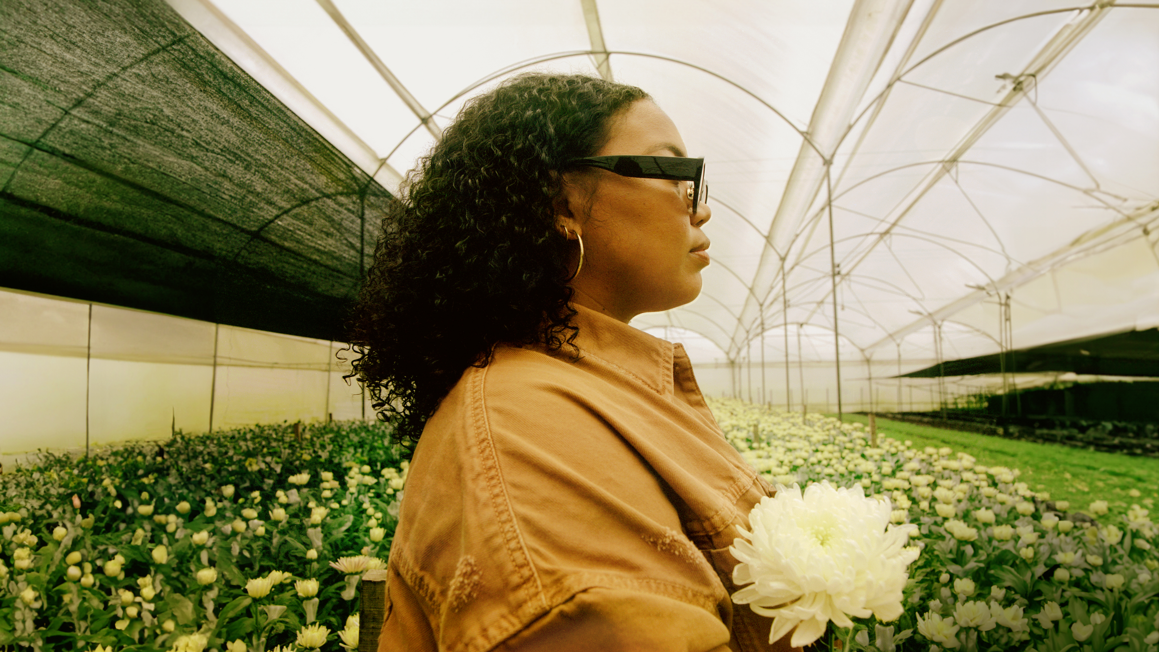 A woman wearing glasses stands in profile, holding a flower in a greenhouse filled with white flowers