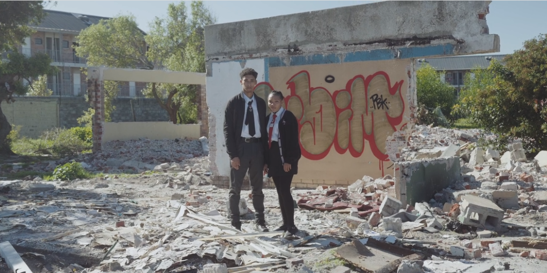 Two teenagers, a boy and a girl, stand on the rubble of a construction. They both wear a white shirt, a tie, a dark jacket, and pants.