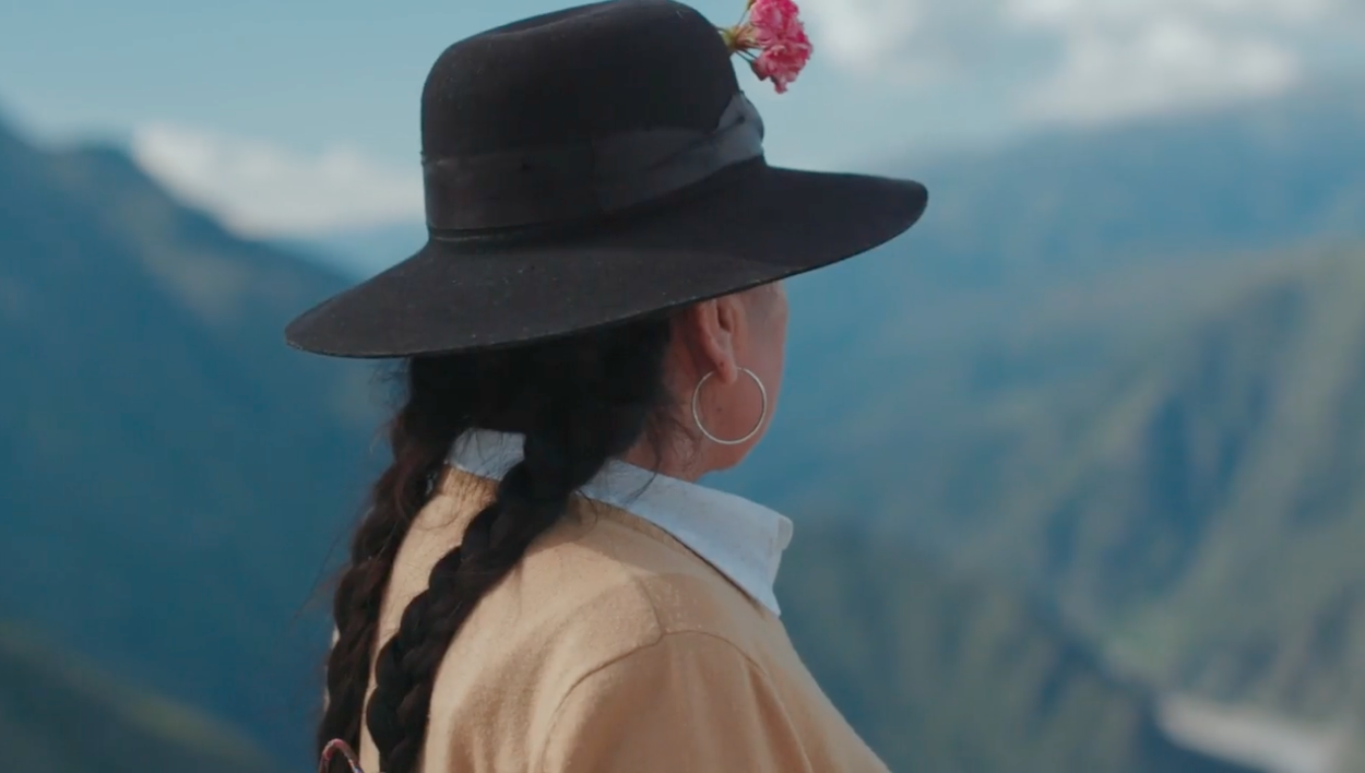 A woman wearing a black hat, a white shirt, and a yellow sweater looks toward a mountain chain in the background. She wears her black hair in two braids.