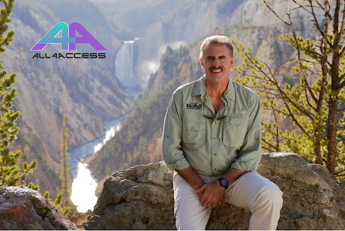 A man in his 50s seats on a big rock. His khaki shirt has embroidered the phrase Mundo salvaje con Ron Magill. On a deep canyon behind him, a white river flows. The All4Access logo appears on the top left of the image.
