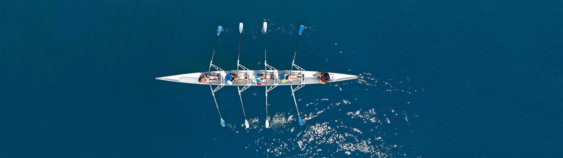On the blue water, 4 people row in sync in a long, thin boat.