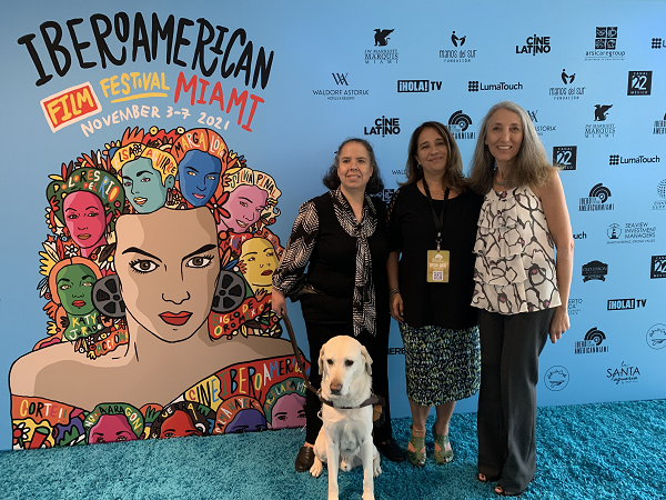 Three women stand in front of an Iberoamerican Film Festival Miami mural. A guide dog sits in front of the woman on the left.